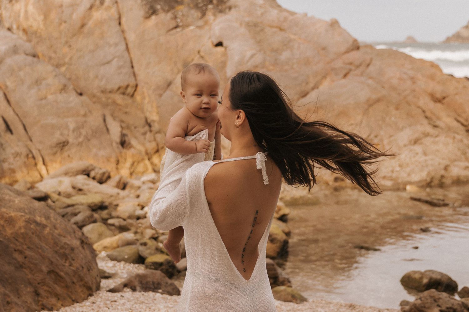 Intimate beachside photoshoot featuring white flowy clothing against rocky coastal backdrop.