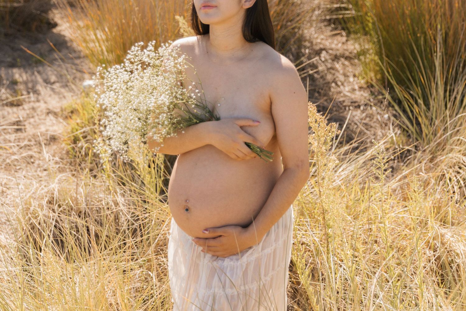 Artistic maternity photo in golden field with white flowers and flowing dress at sunset.