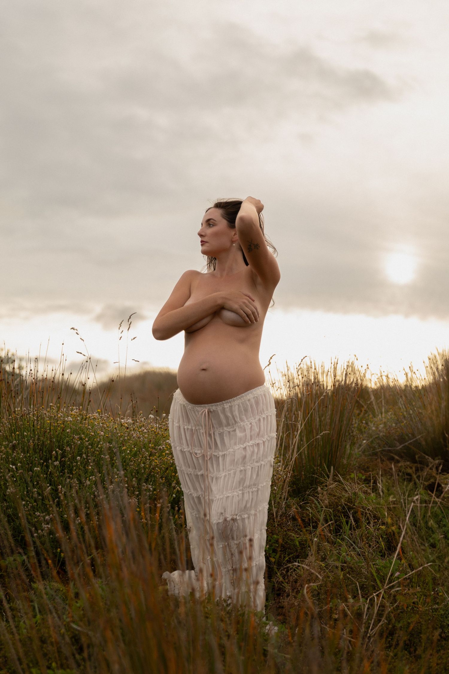 Ethereal maternity photography against cloudy sky and tall grasses at golden hour.