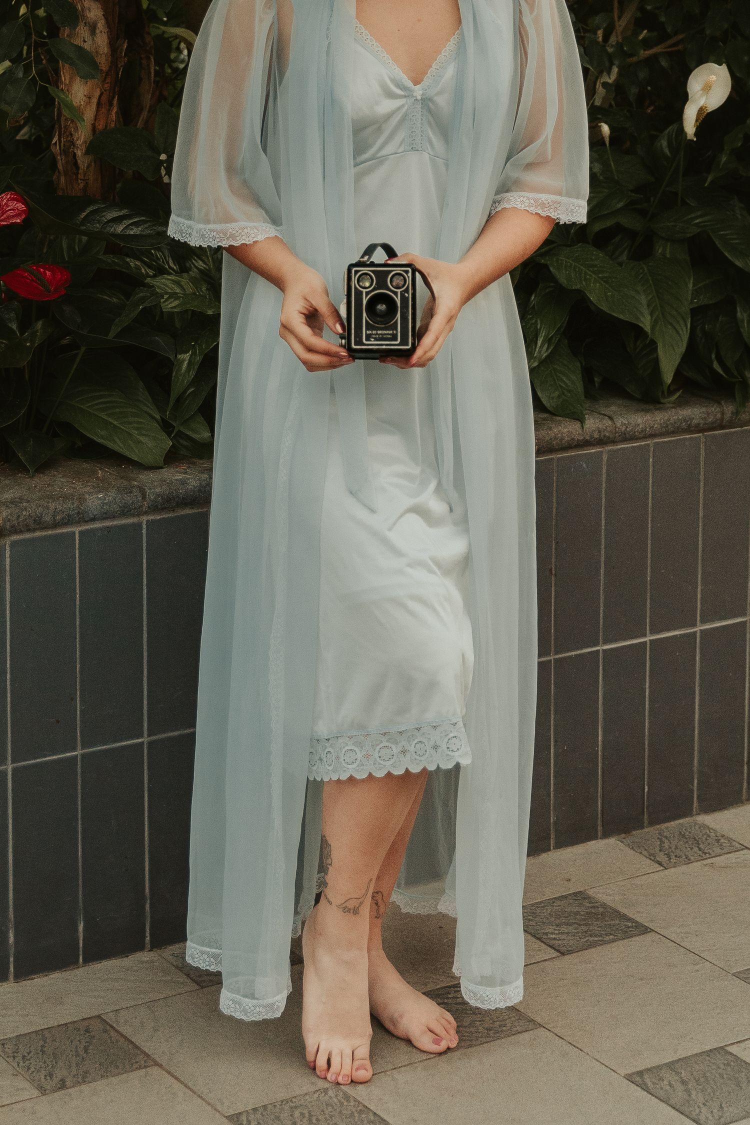 Person in light blue vintage dress holding vintage camera on tiled patio.