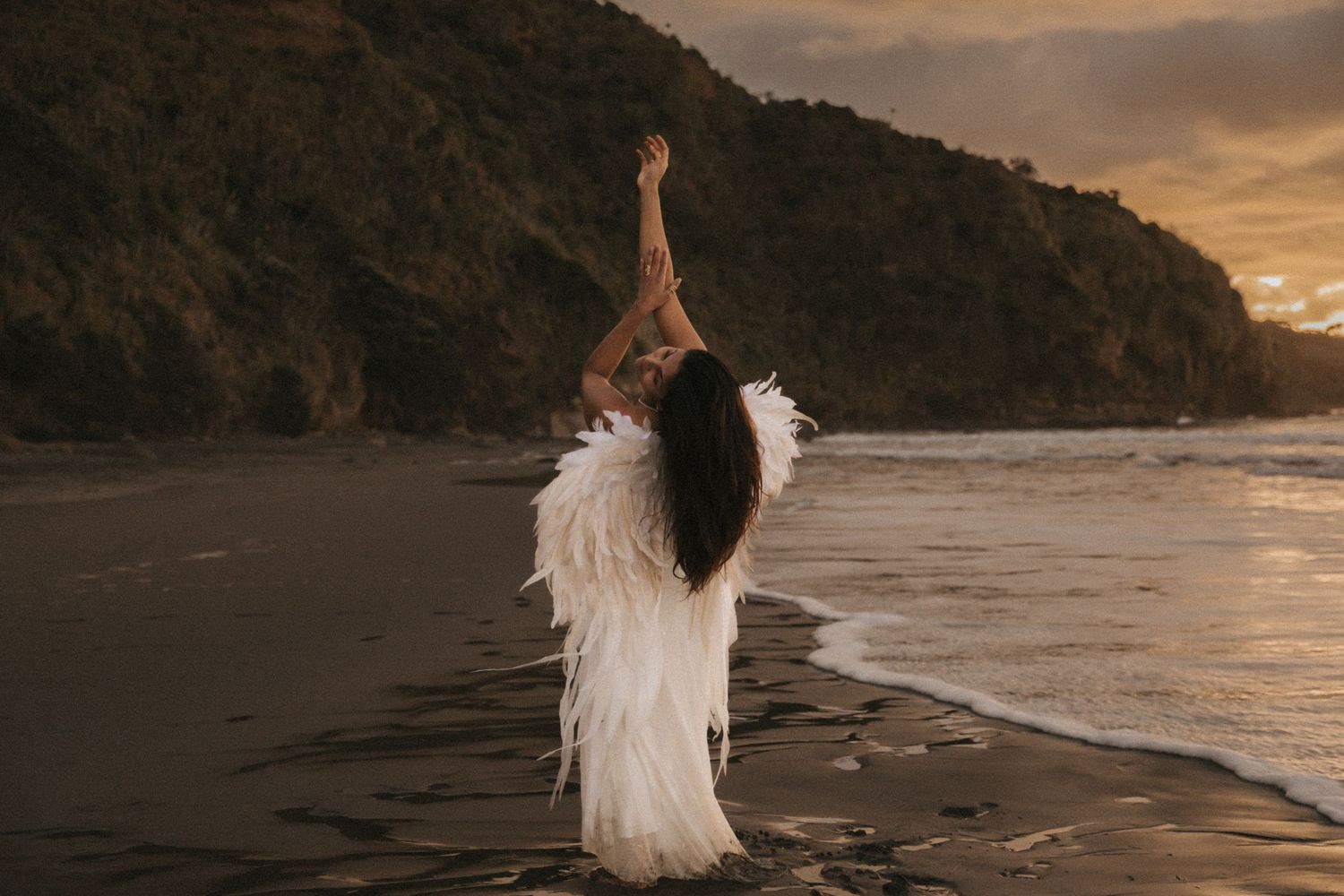 A figure in a flowing white dress walks along a scenic beach at sunset with mountains in the background.