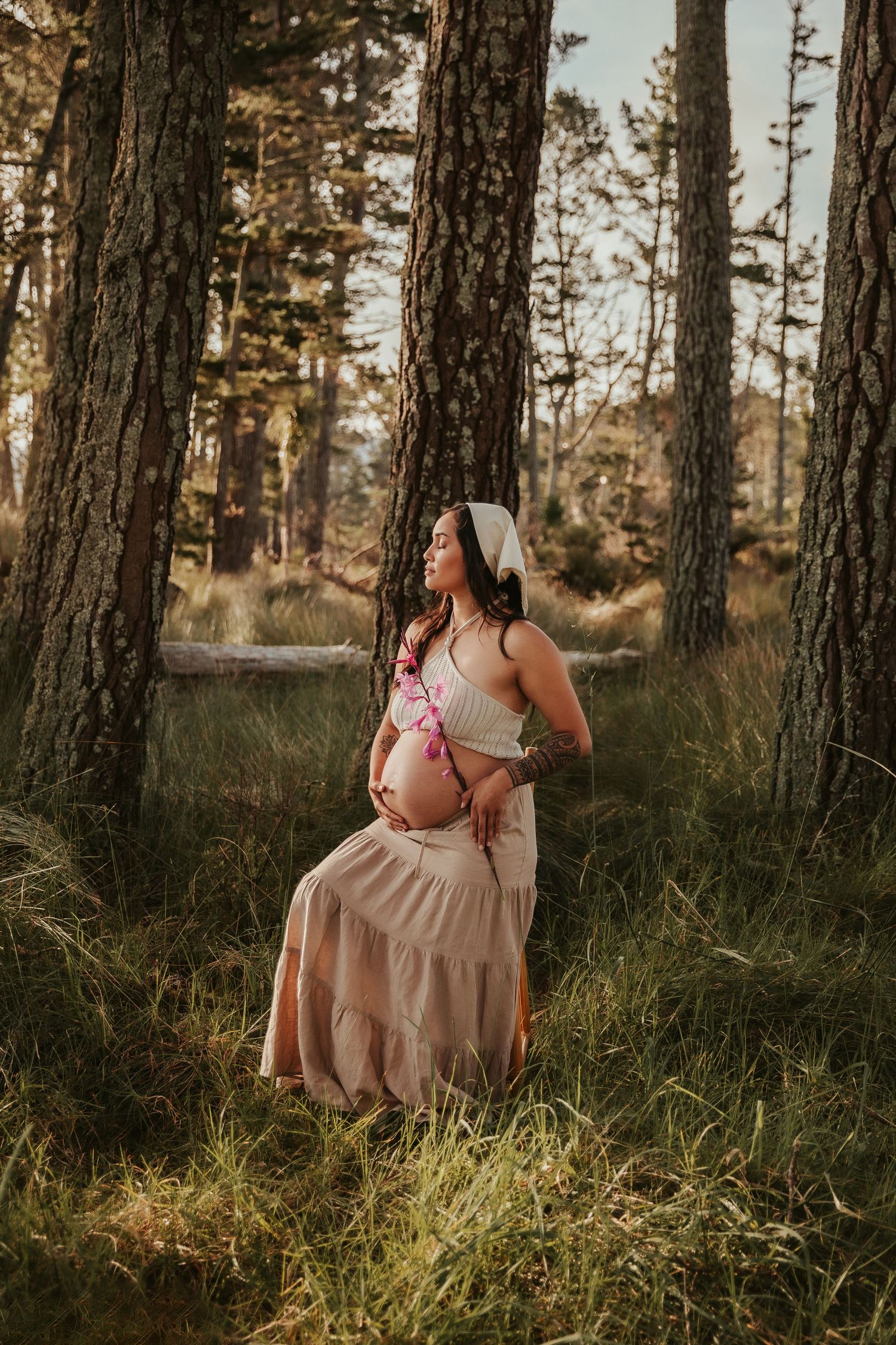 Person in a flowing beige dress poses gracefully among tall pine trees in golden sunlight.