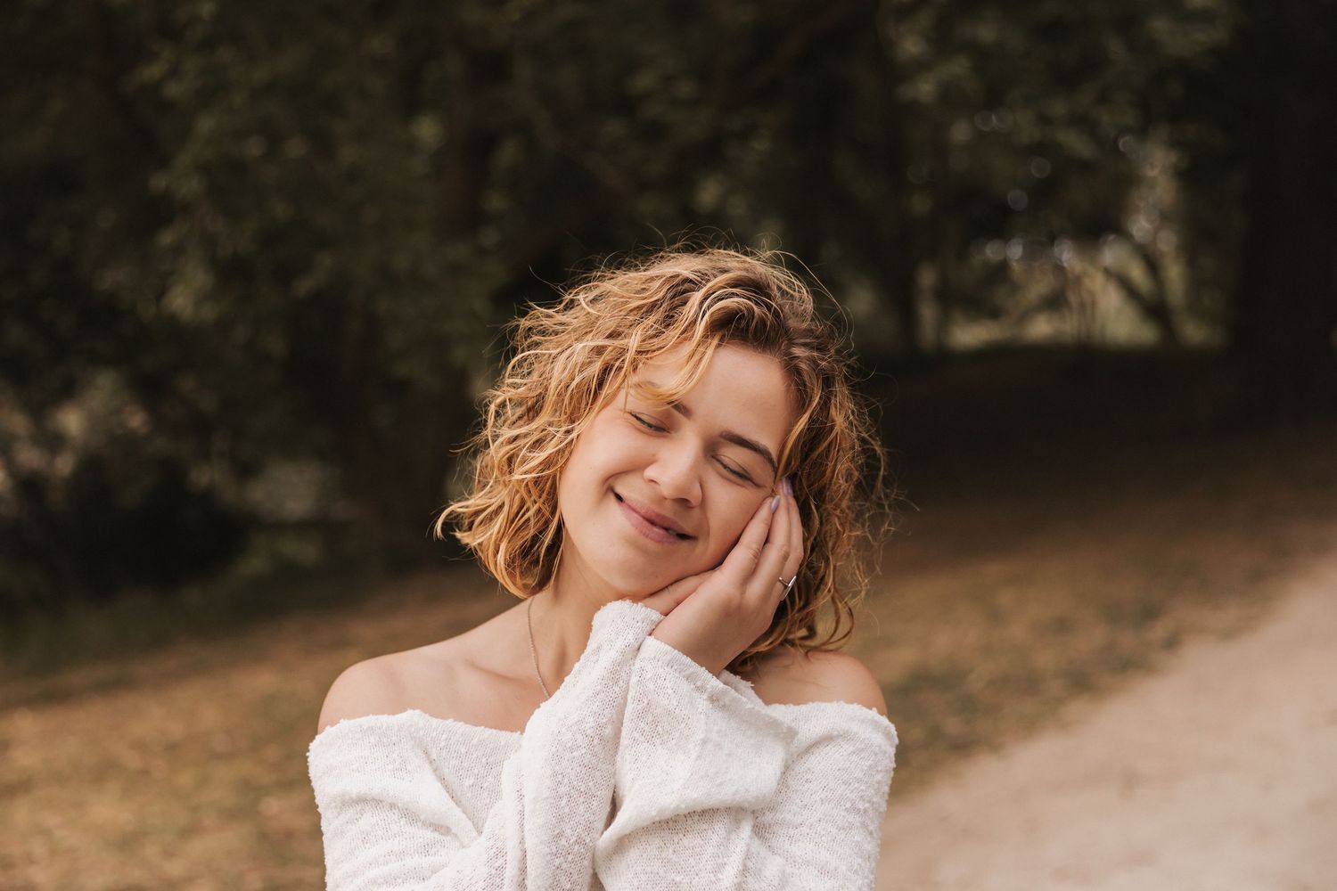 Woman in white sweater poses with hand on cheek and closed eyes outdoors.