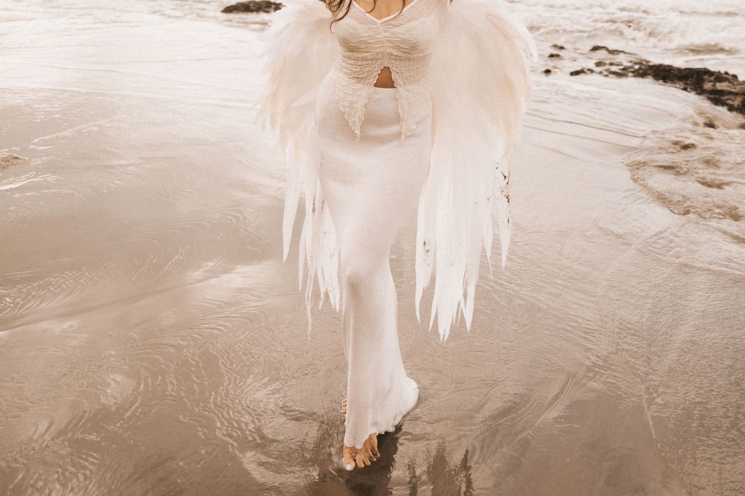 Flowing white dress and bare feet walking along wet sand on a beach in warm sepia tones.