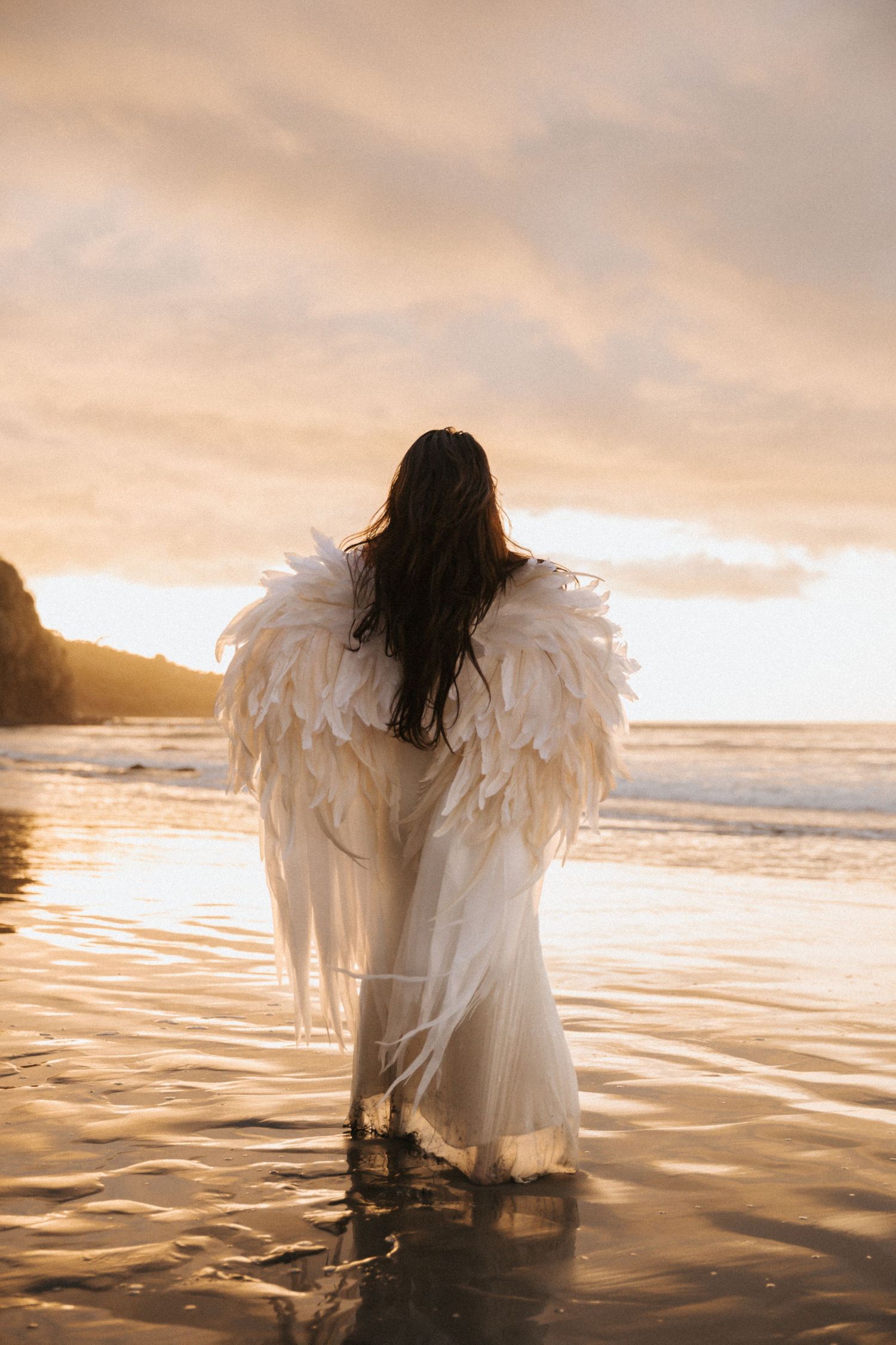 A figure in white angel wings sits peacefully at the ocean's edge during a golden sunset.