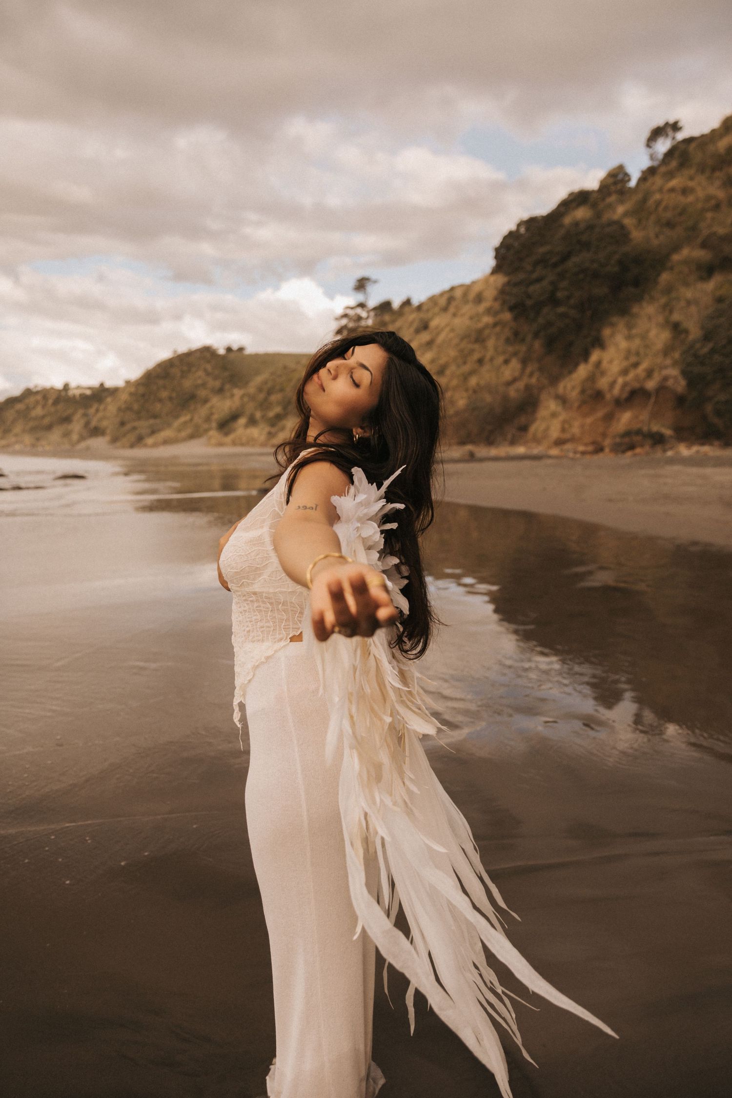 A figure in a flowing white dress poses dramatically on a moody beach with cliffs in the background.