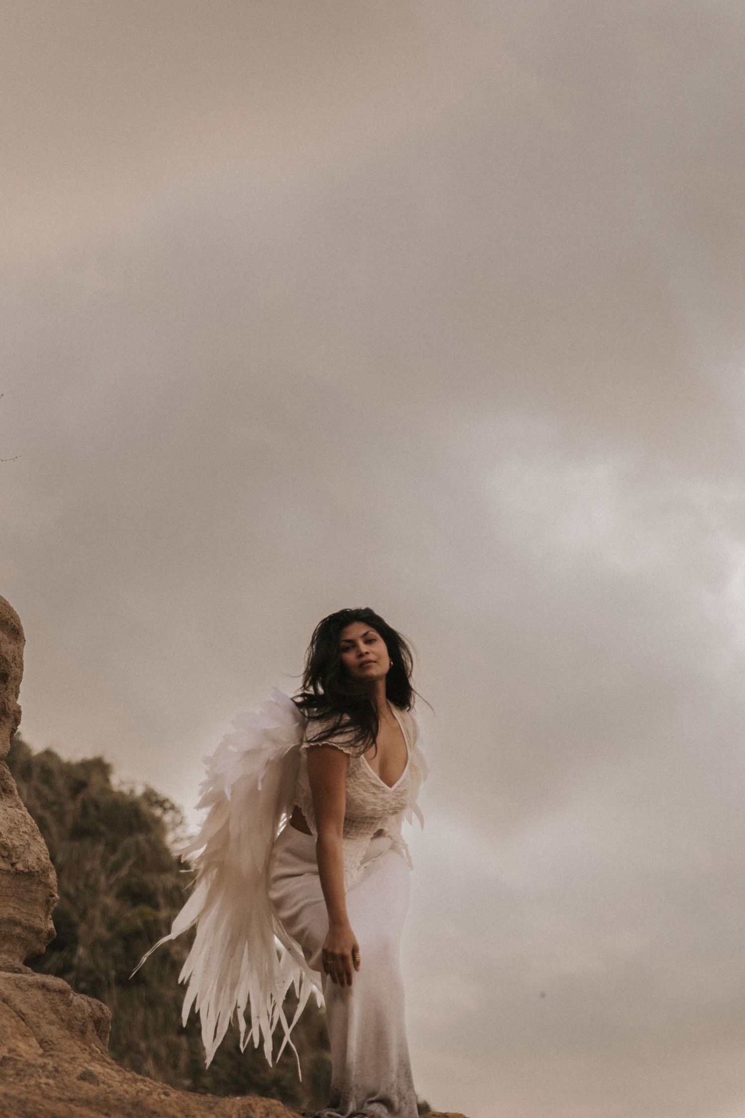 A figure in a flowing white dress dances ethereally against a moody cloudy sky on a rocky terrain.