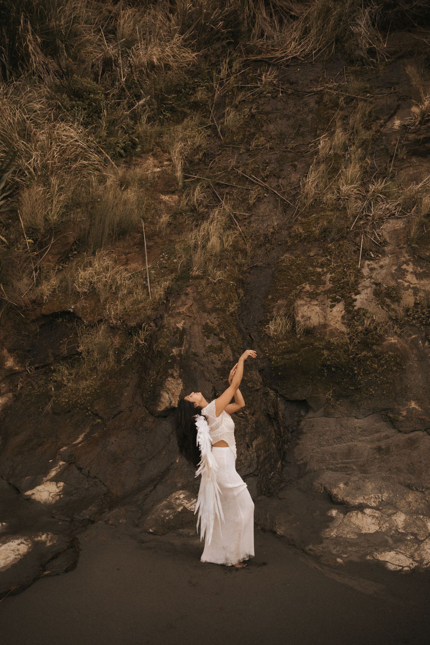 A dancer in white performs graceful movements along a rugged cave wall in dramatic lighting.