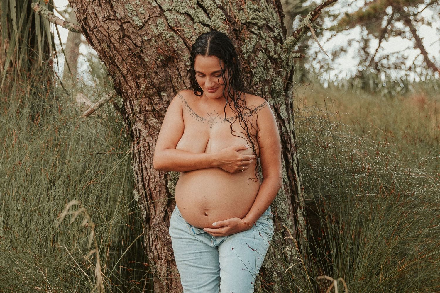 Pregnant woman in light blue jeans poses in a natural outdoor setting with tall grass and trees in the background.