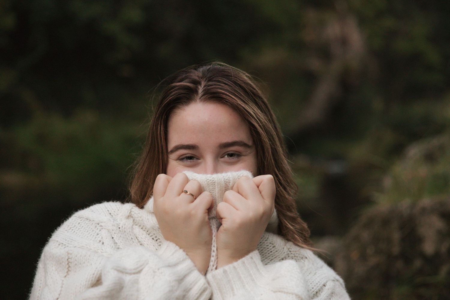 Woman in cozy white knit sweater covering half of her face with the collar on a moody autumn day with a cheeky smile.