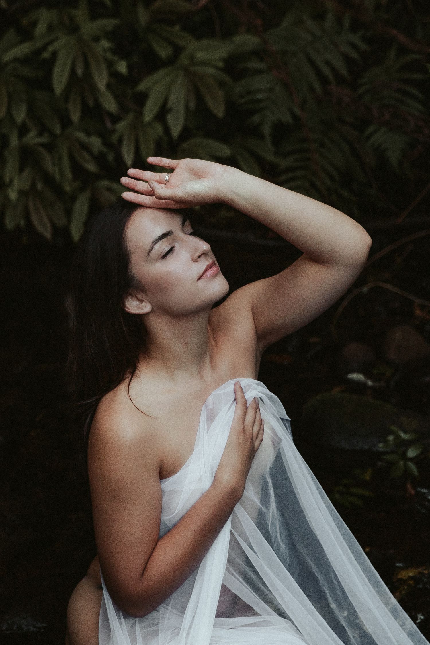 A dreamy portrait with sheer white fabric and dark foliage creating a moody artistic atmosphere.