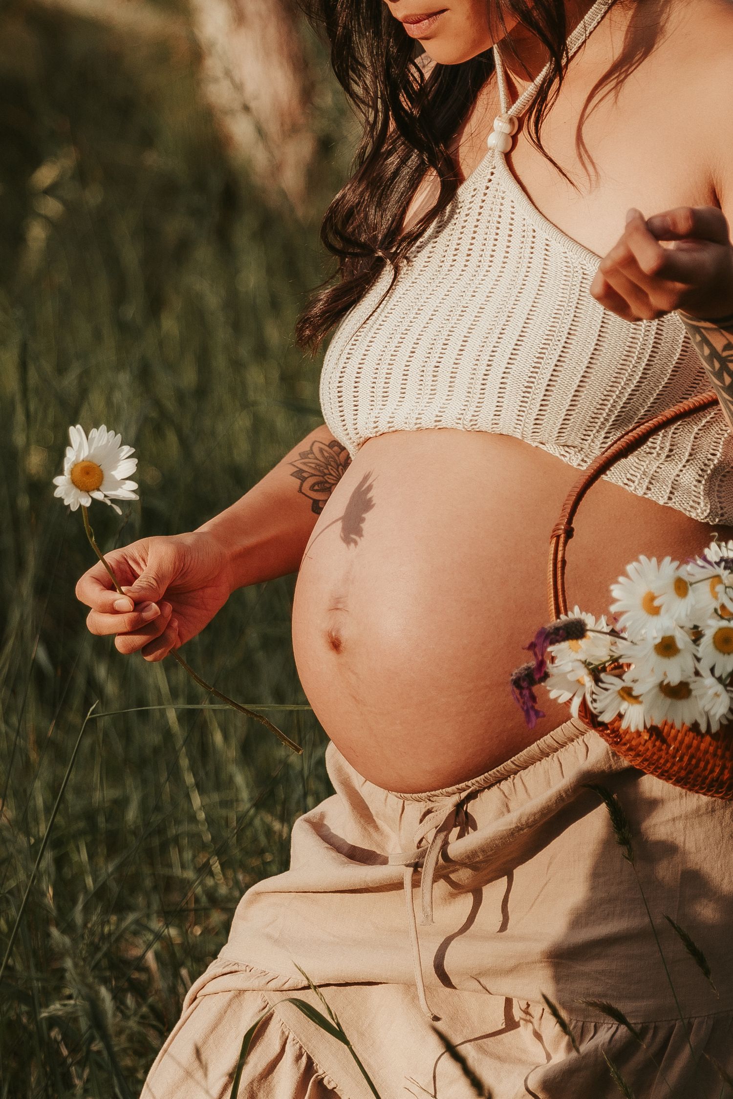 A pregnant belly in a cream knit top is shown with delicate daisies against a natural outdoor setting at sunset.