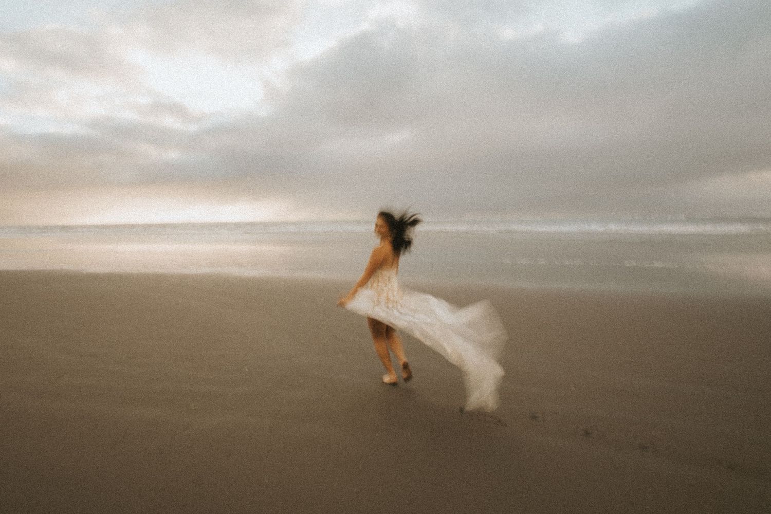 A flowing white dress catches the ocean breeze on a cloudy beach.