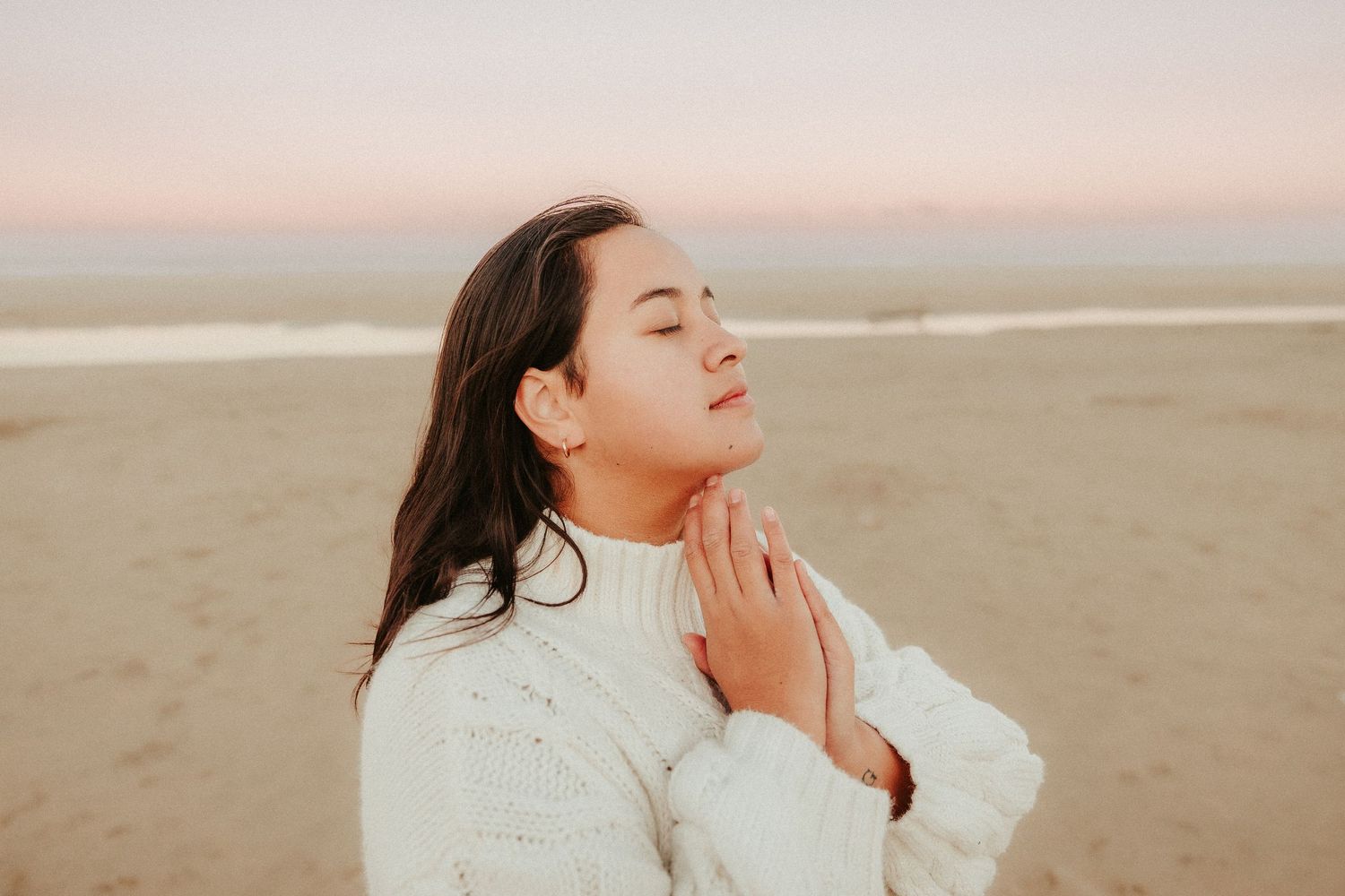 A woman in a white sweater enjoys a peaceful moment at the beach during sunset.