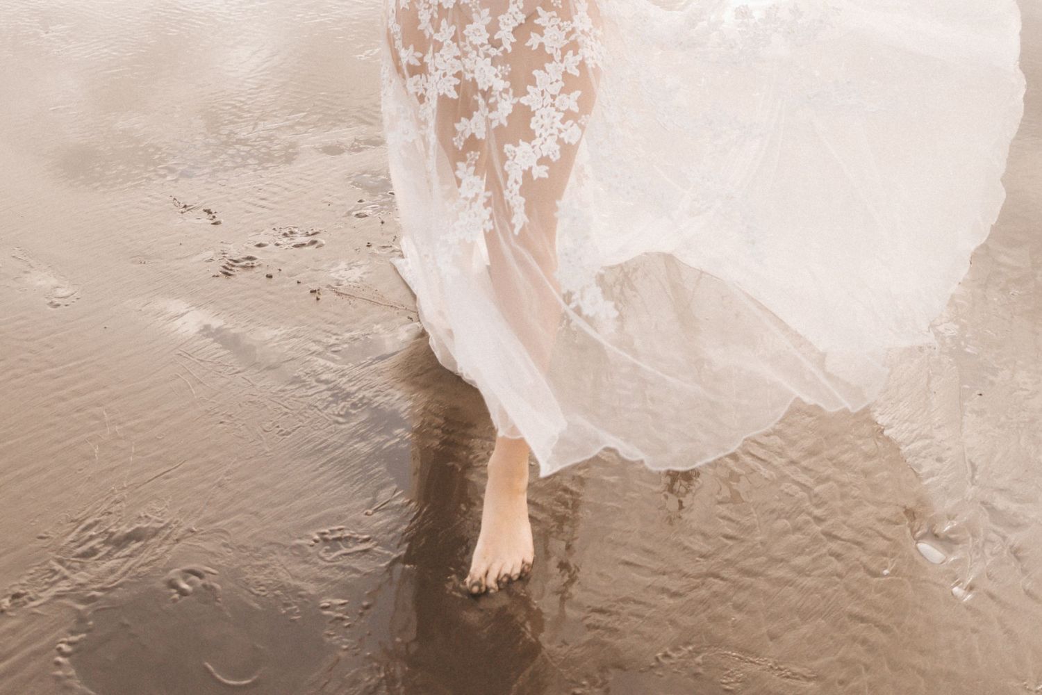 Close-up detail of delicate white lace wedding dress against sandy beach background.