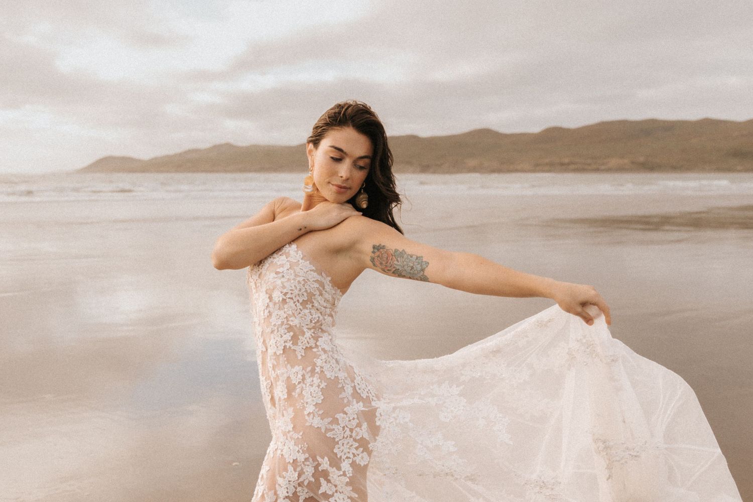 Ethereal beach photoshoot featuring a delicate white lace dress flowing in the wind against a moody coastal backdrop.