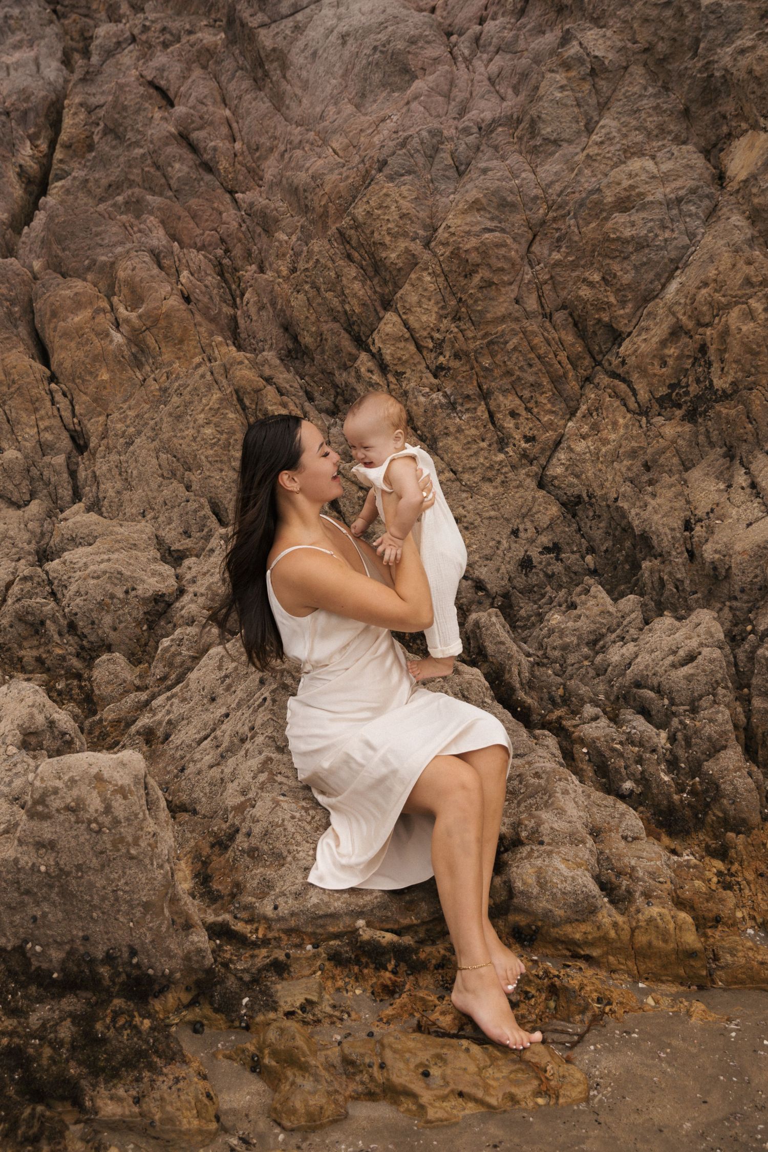 A tender moment captured against a rocky cliff backdrop with a flowing white dress creating an ethereal atmosphere.