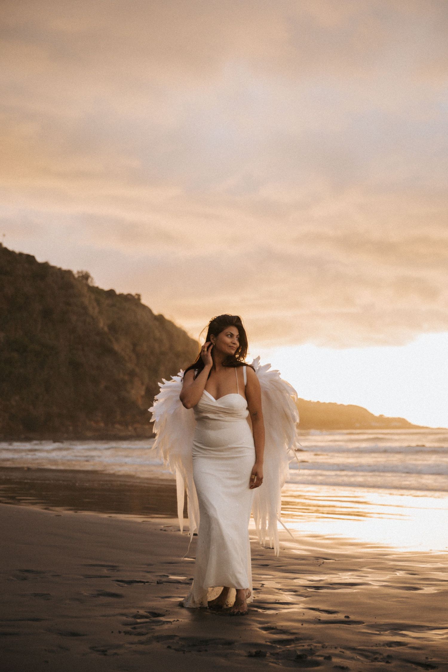 Figure in white dress with angel wings poses gracefully on beach at golden sunset.