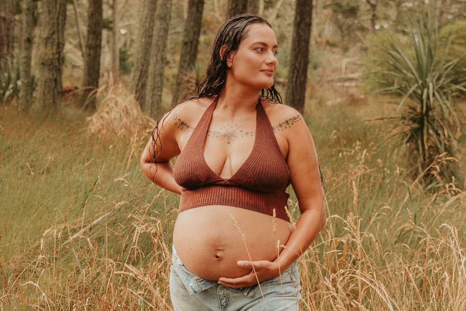 A pregnant woman  poses outdoors in a natural setting with tall grass and trees.