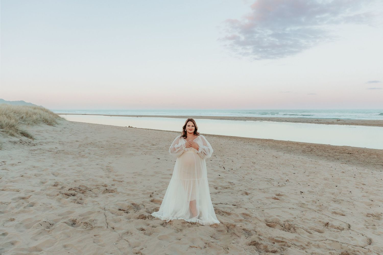 A pregnant woman in a flowing white dress walks along a sandy beach at sunset with waves crashing in the background.