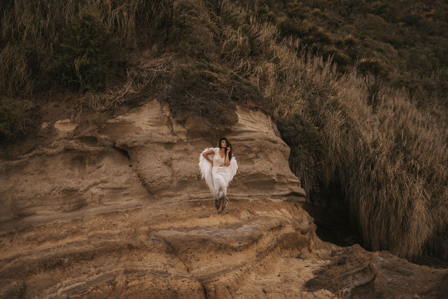 A couple in wedding attire poses on a rocky cliff edge overlooking a dramatic mountain landscape at sunset.