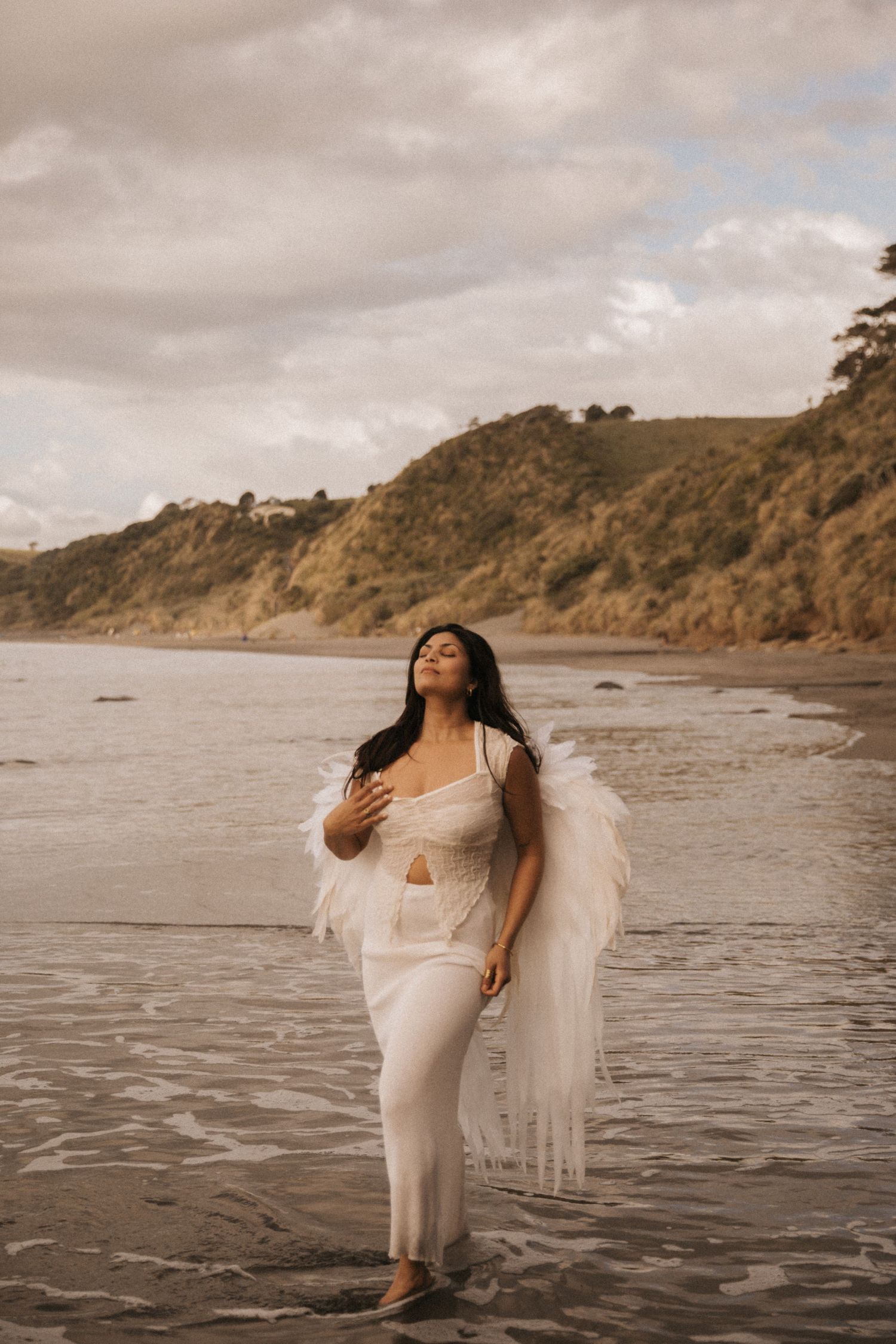 A person in a flowing white dress poses gracefully on a moody beach with dramatic cliffs in the background.