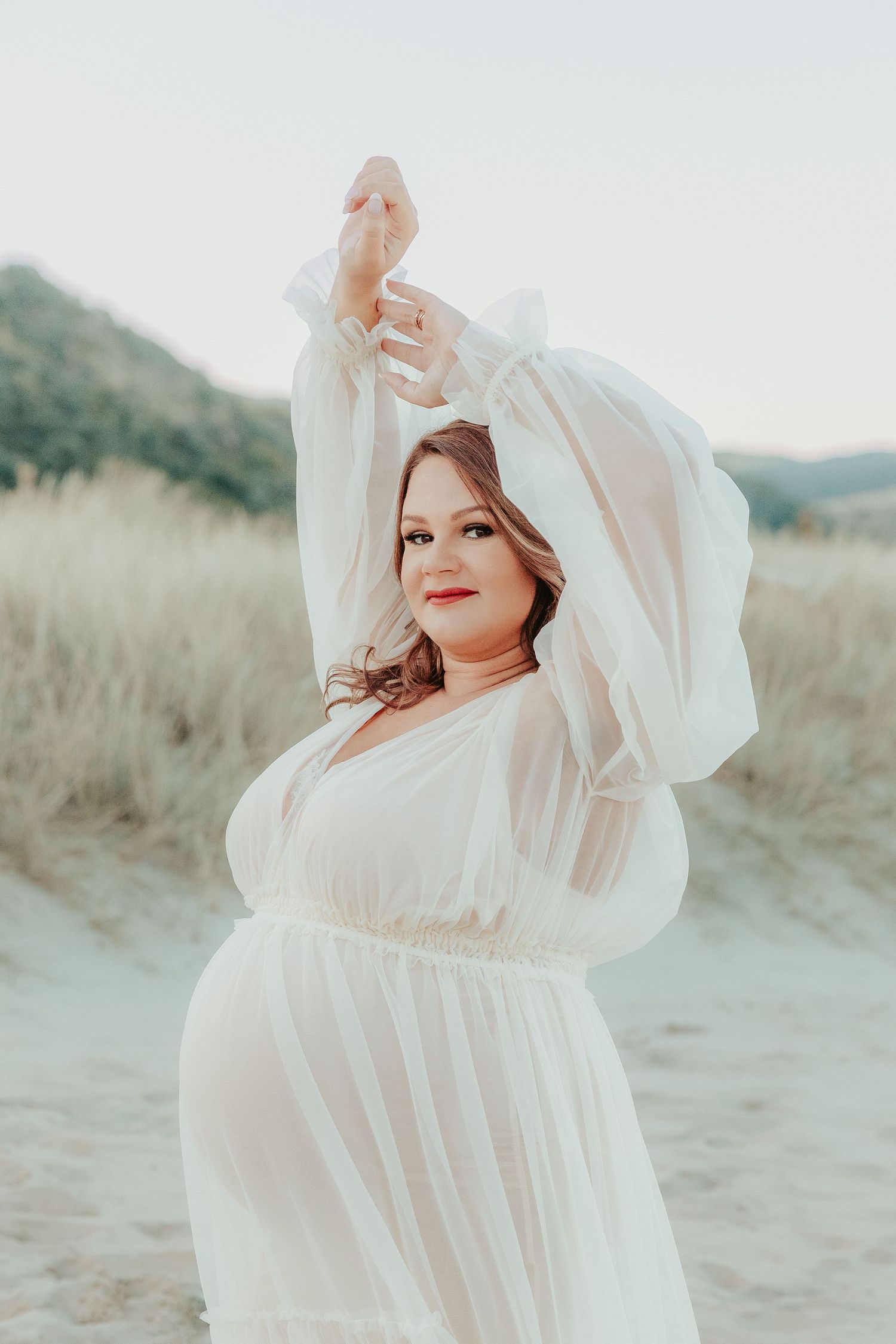 A pregnant woman in a flowing white dress poses gracefully against a soft beige beach landscape at sunset.