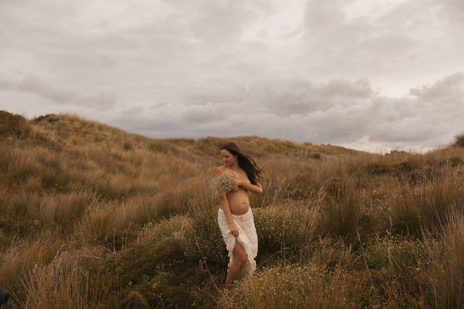Dramatic landscape maternity photography with cloudy sky and rolling hills.