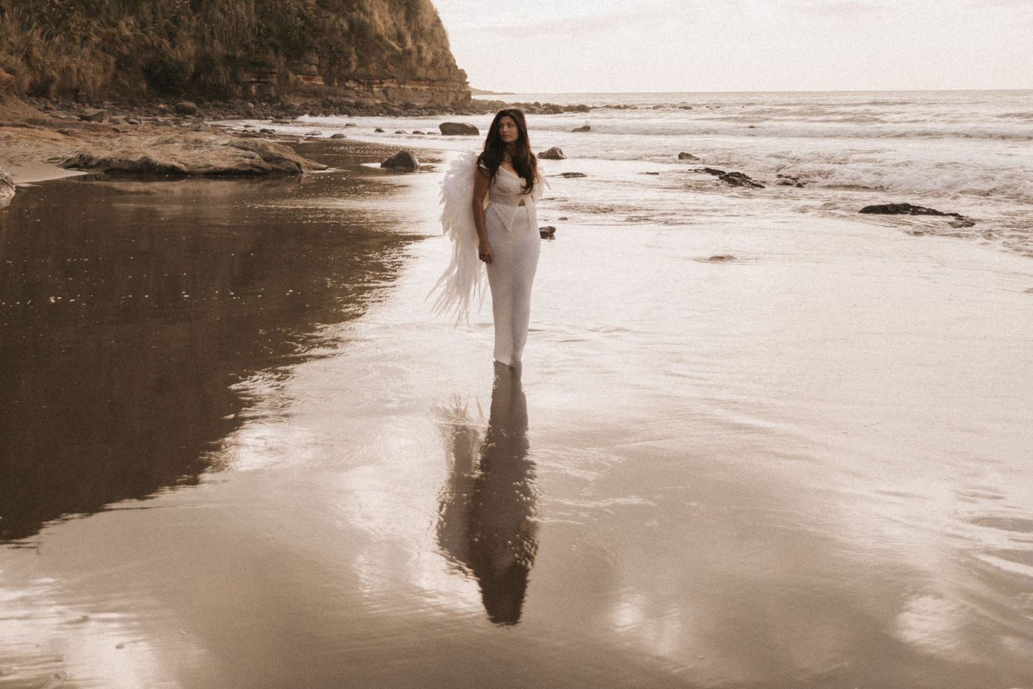Person in white dress walking along misty beach shoreline at sunset creating reflections in wet sand.