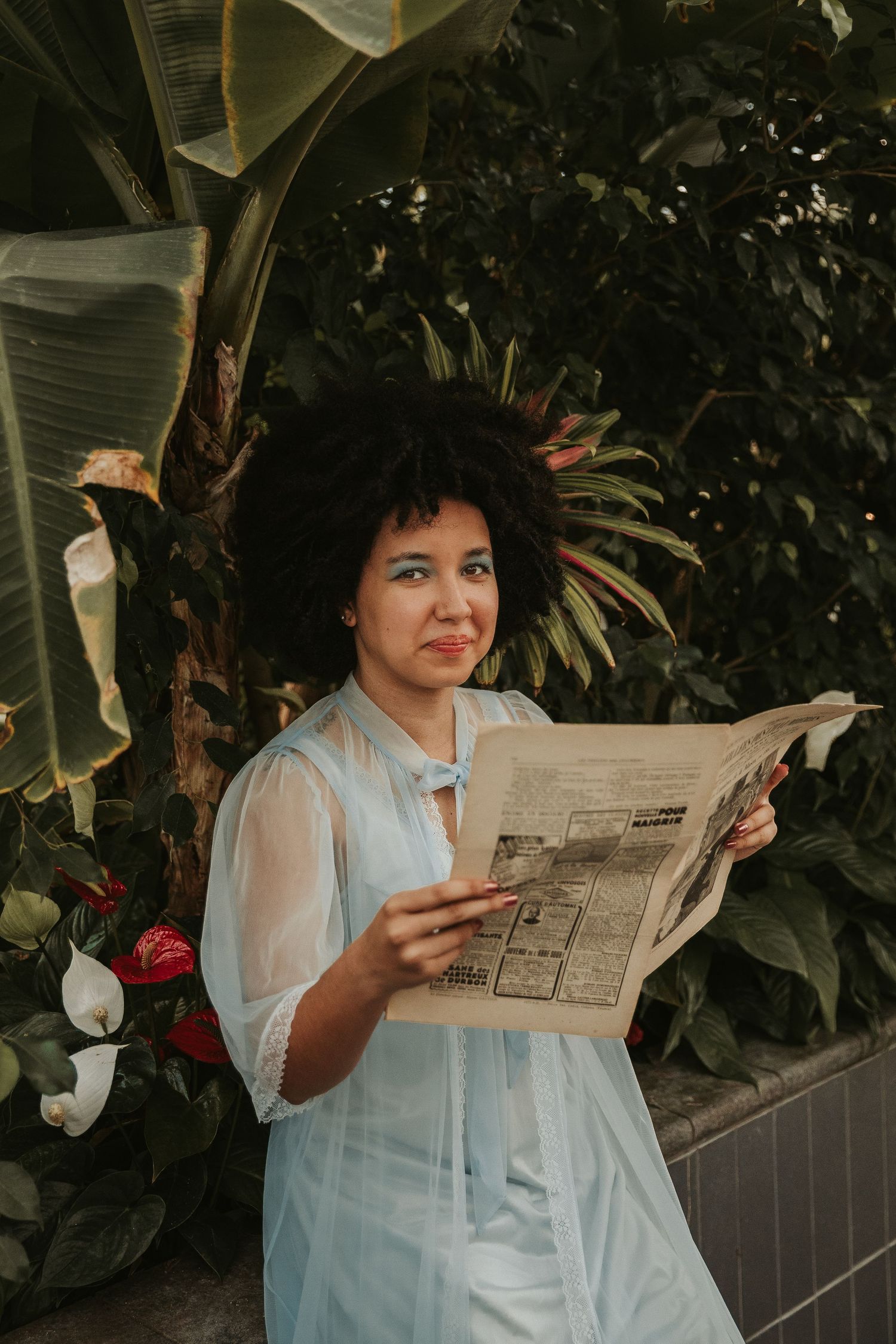 Woman in light blue vintage dress reading newspaper under tropical foliage.