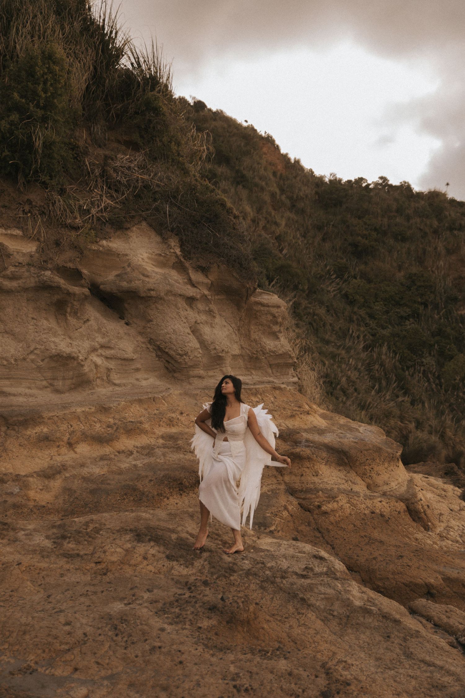 A couple in white attire poses dramatically on a rugged cliffside during a sepia-toned photoshoot.