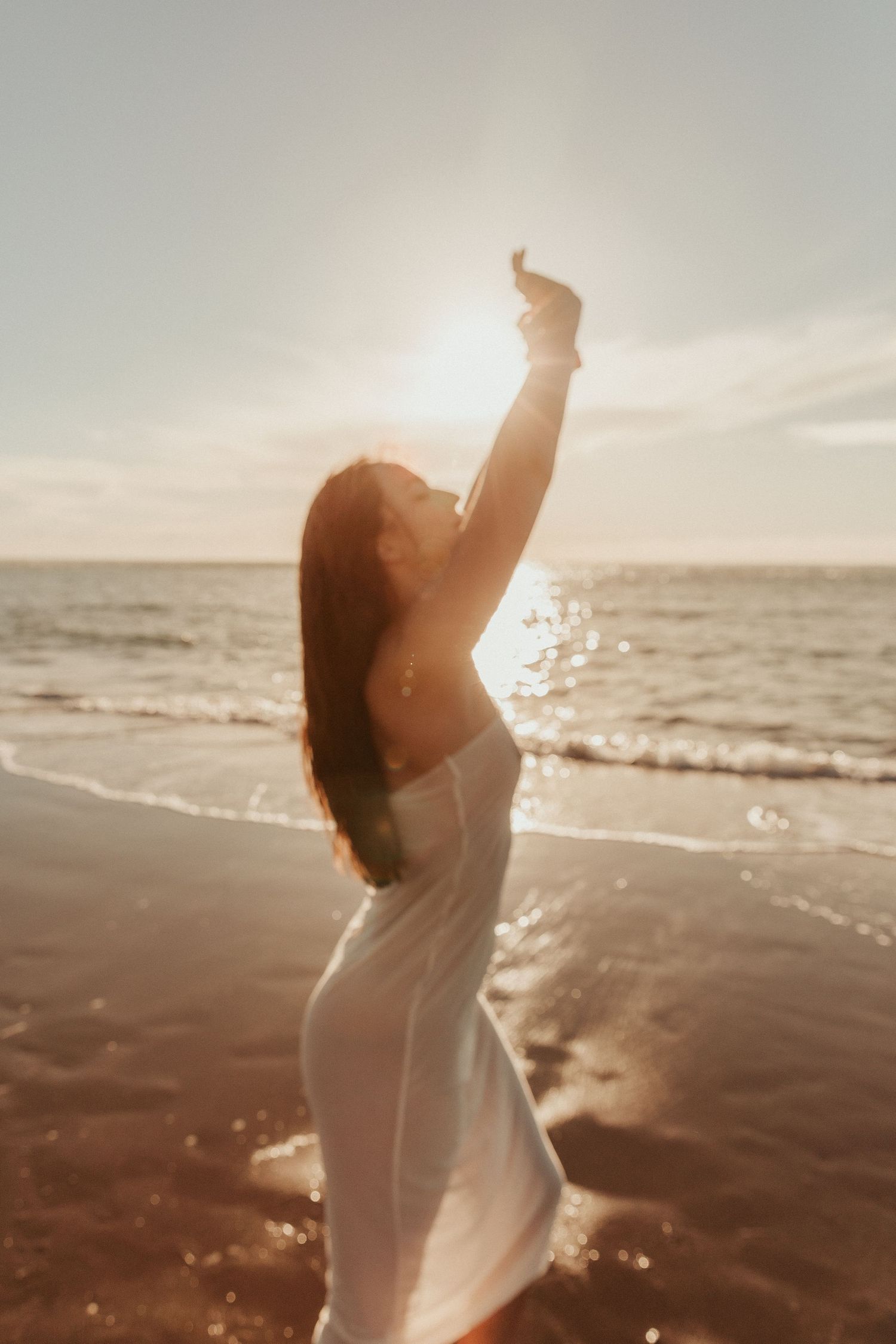 Silhouetted figure in flowing dress raises arms toward sunset over ocean waves.