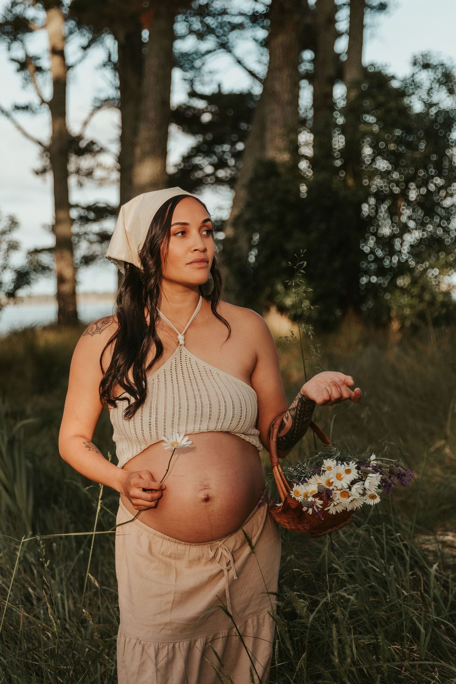 A maternity photoshoot in a grassy field at sunset shows a woman in a beige dress holding her pregnant belly.