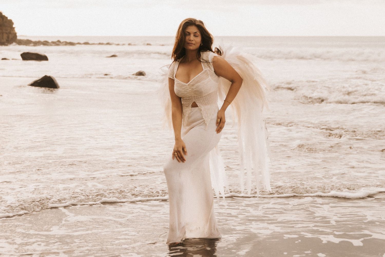 A figure in a flowing white dress walks along a misty beach shoreline at sunset.