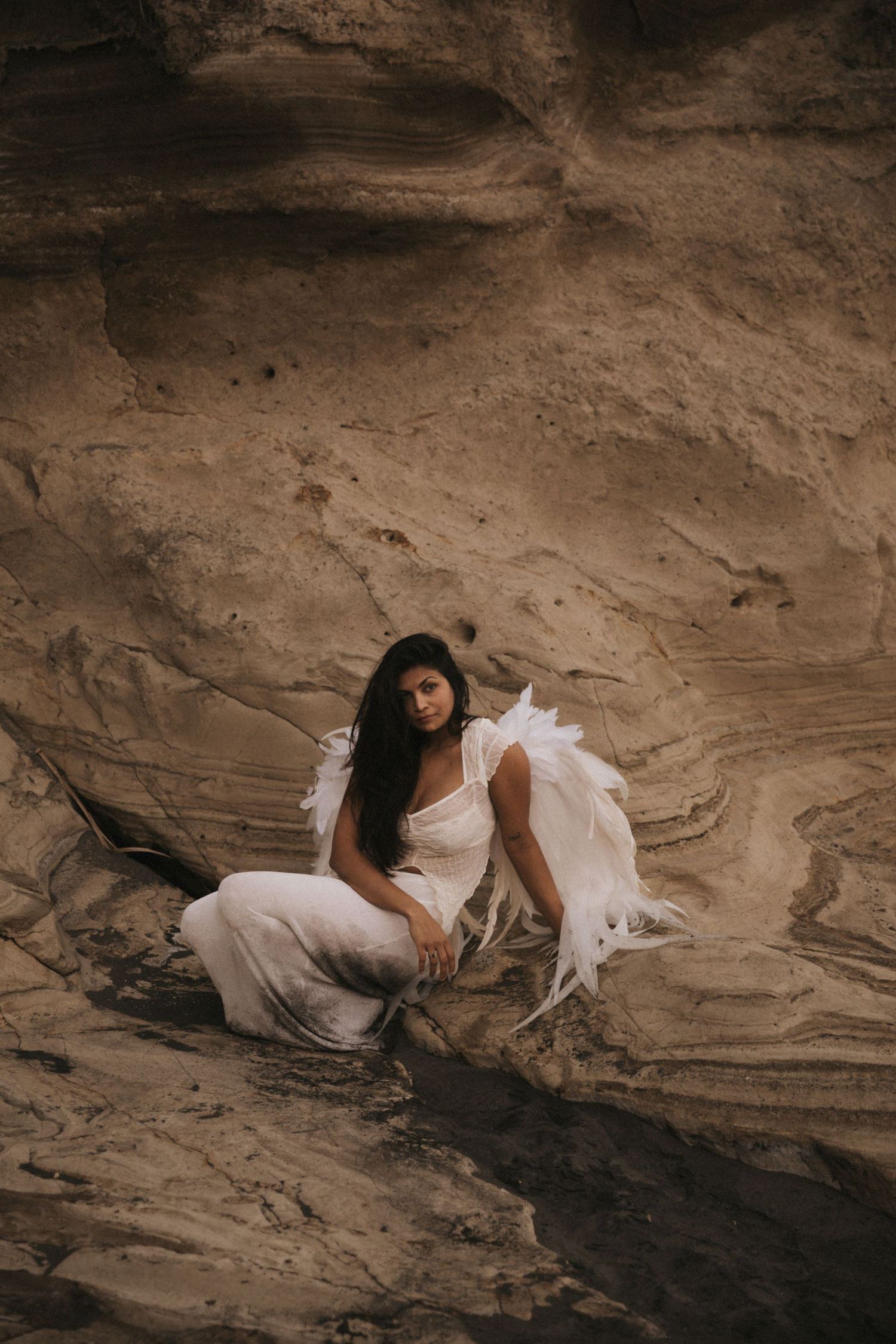 A person in a white dress reclines artistically against a textured rock formation in a moody sepia-toned photoshoot.