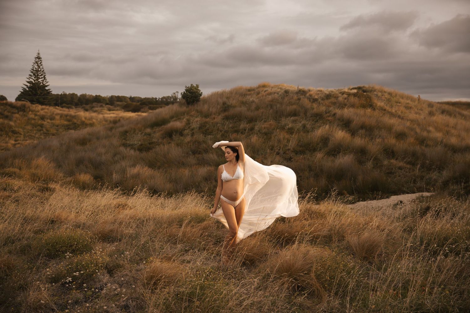 Ethereal maternity photoshoot in a golden field at sunset with flowing fabric catching the breeze.