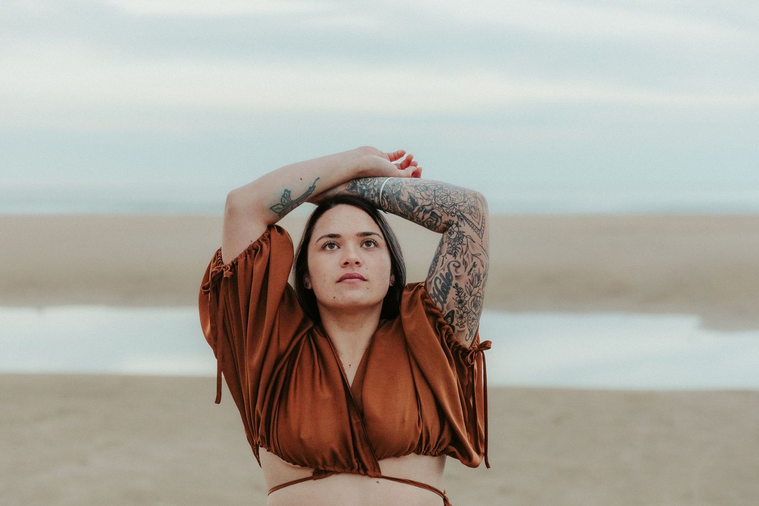 Dramatic beach portrait with flowing rust-colored fabric against cloudy sky.