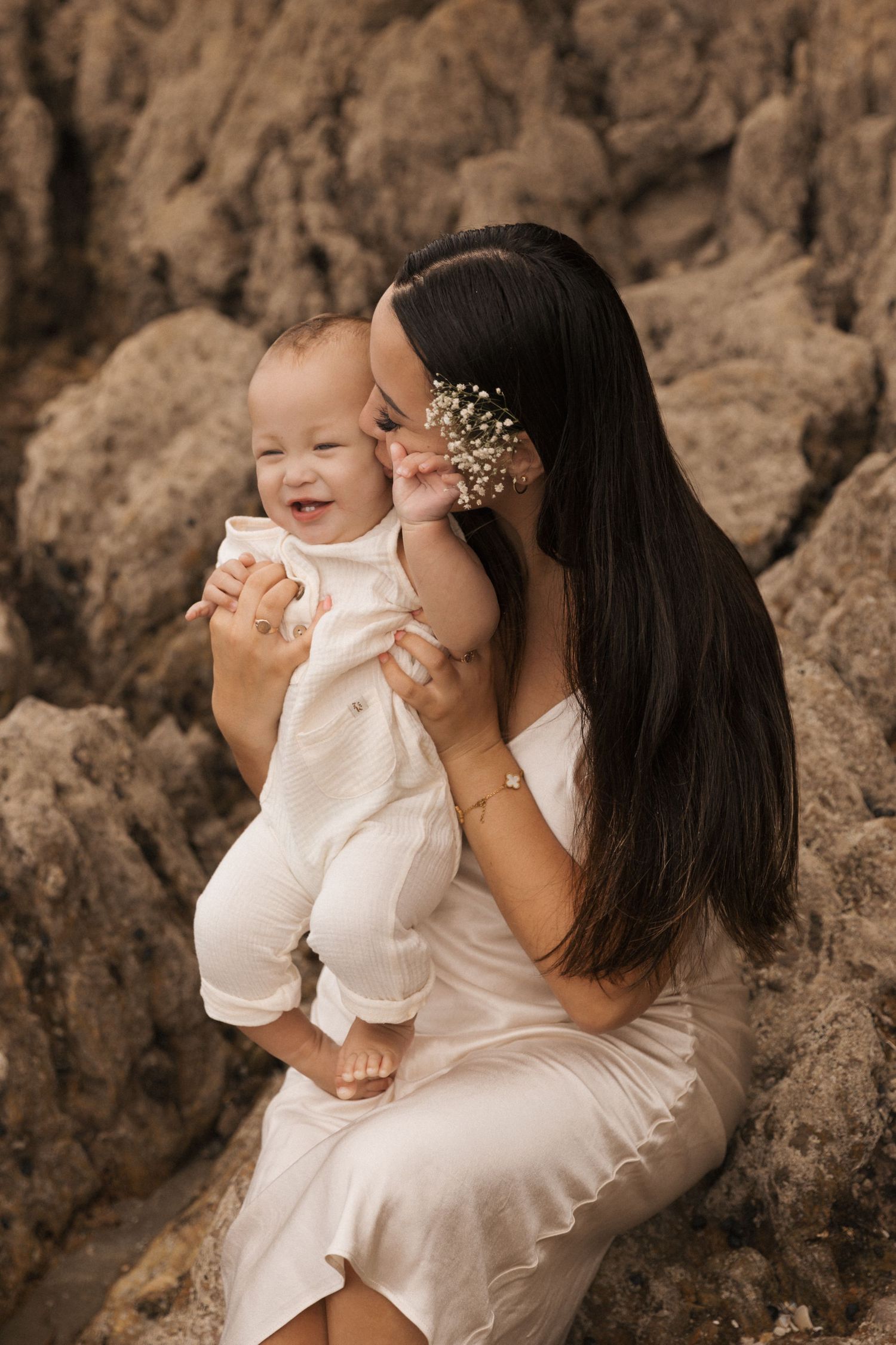 Mother and baby sharing intimate moment against rocky backdrop in white outfits.