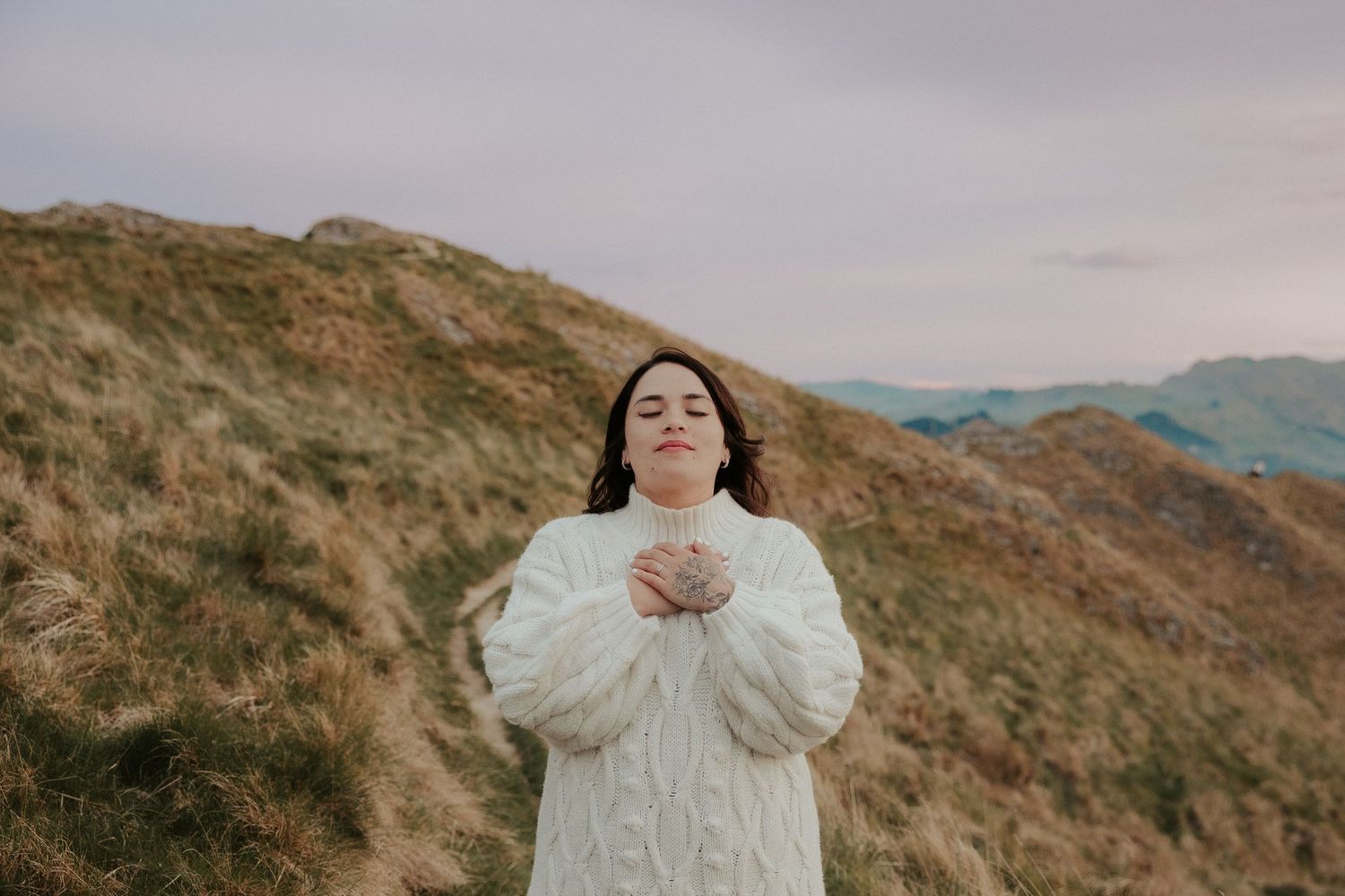 Person in flowing white dress stands gracefully against mountainous landscape.
