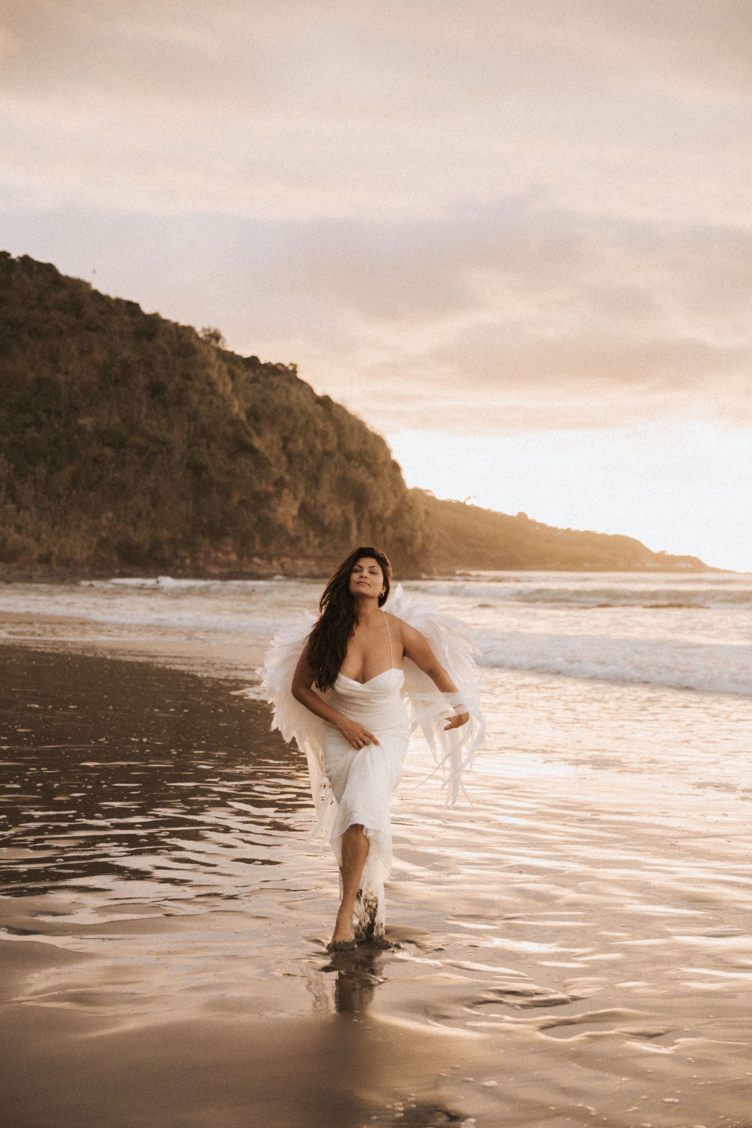 A person in a flowing white dress stands in shallow ocean water at sunset with cliffs and coastline in the background.