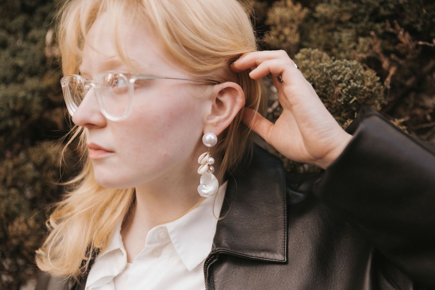 A person with blonde hair and glasses wearing pearl earrings poses against a dark background.