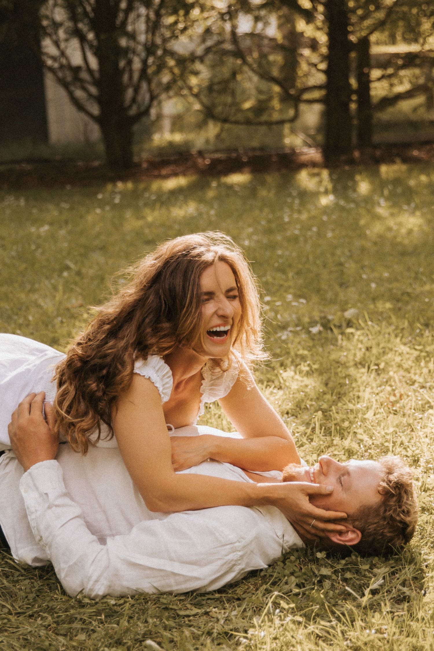 A couple in white clothing lies on grass in sunlight sharing joyful laughter during a romantic outdoor photoshoot.