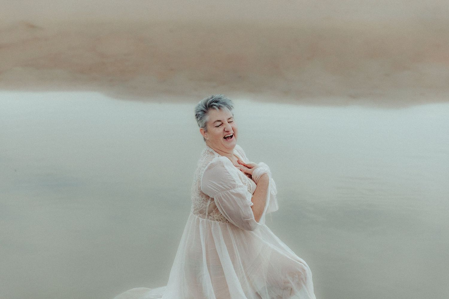 Person in flowing cream dress twirls joyfully in ethereal misty water setting.