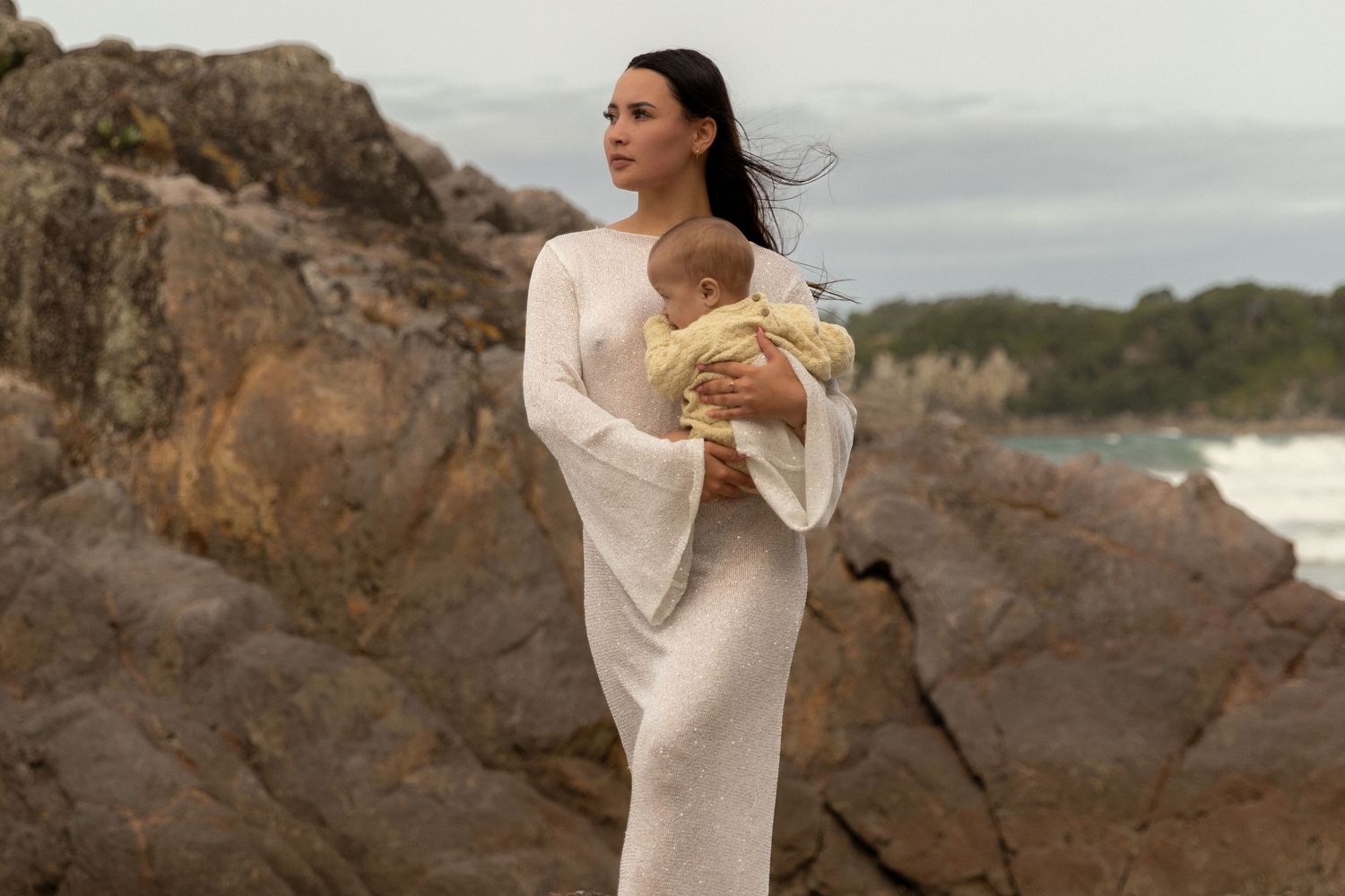 Ethereal beachside photography with flowing white dress against rocky coastline.