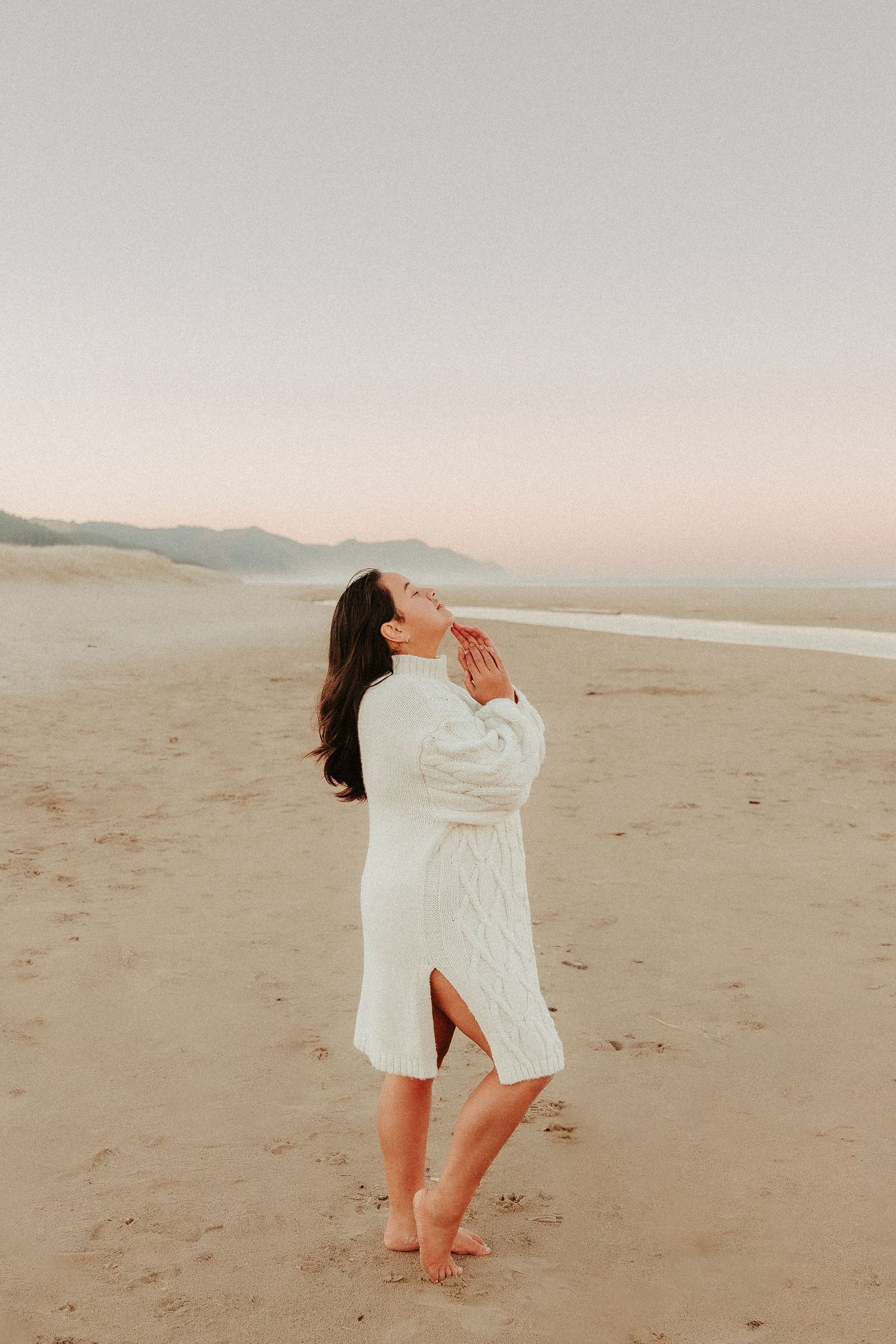 Figure in white clothing dances expressively on a sandy beach at sunset.
