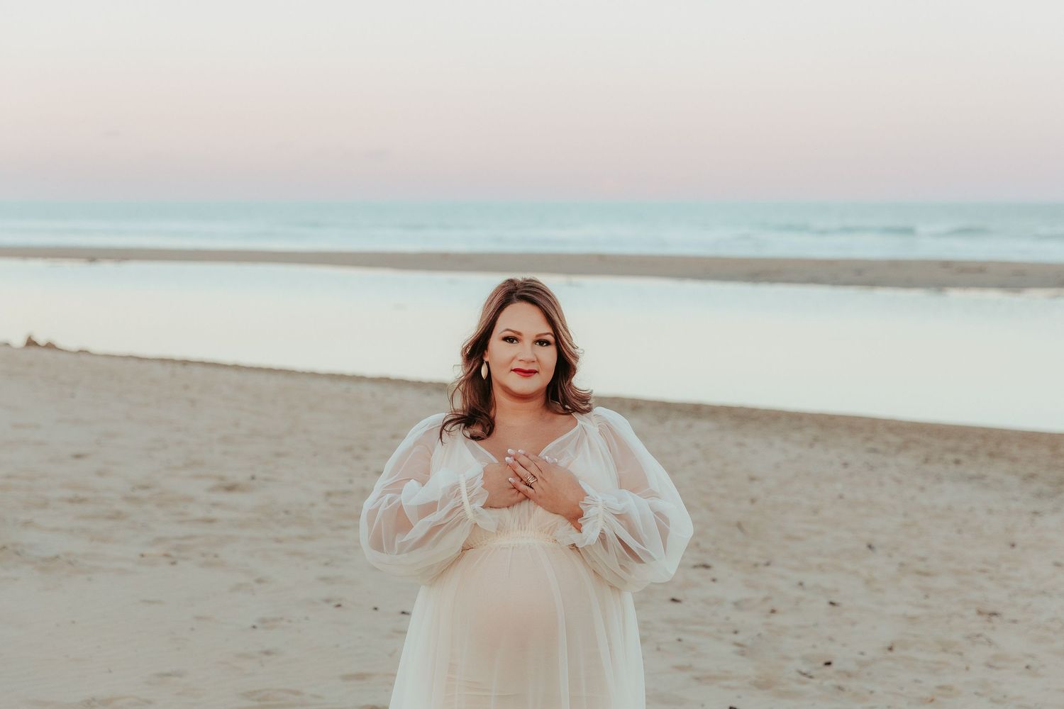 Woman in a flowy white dress walks along a sandy beach at sunset.