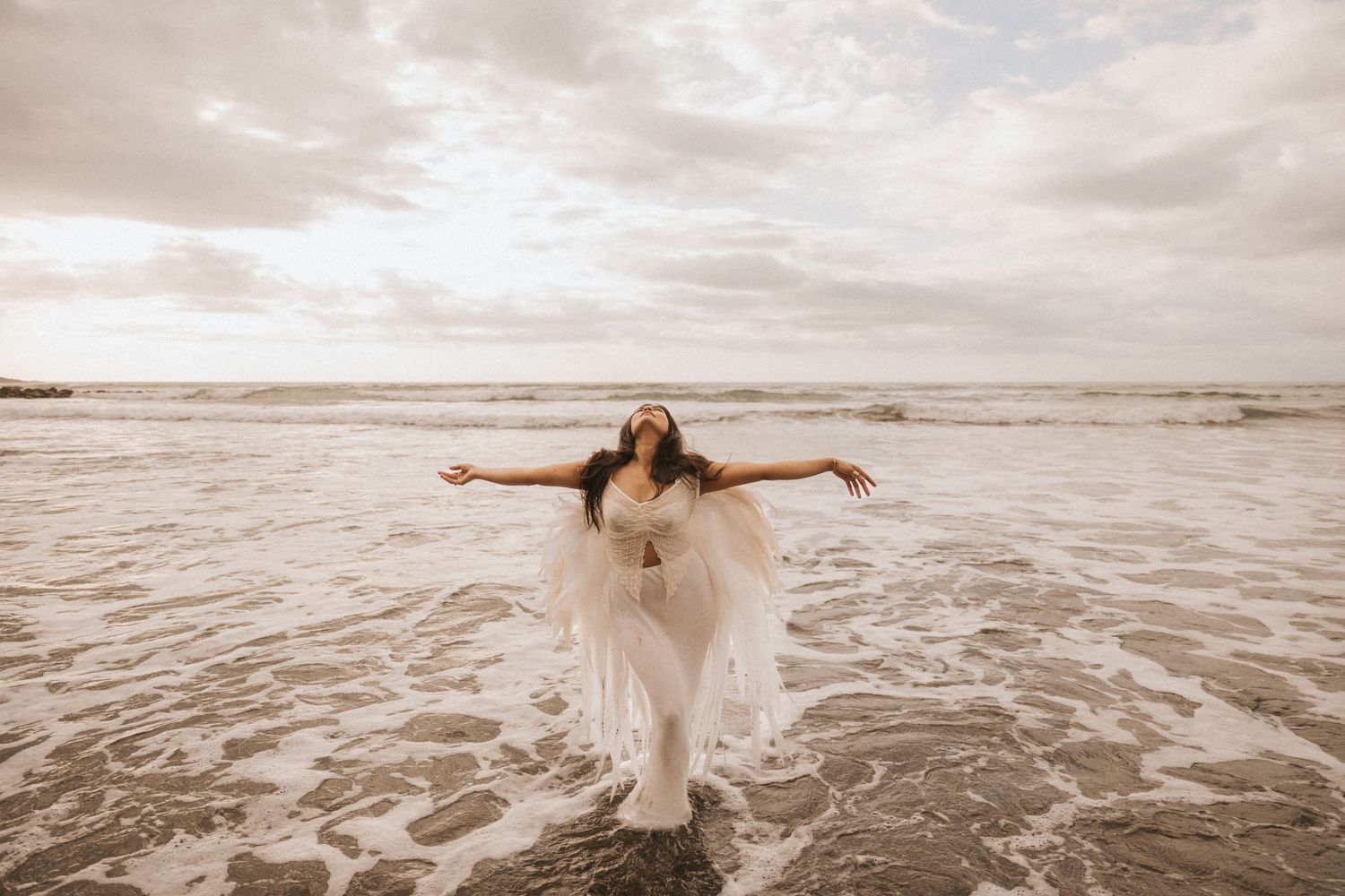 A person in a flowing white dress dances gracefully along the beach shoreline at sunset.