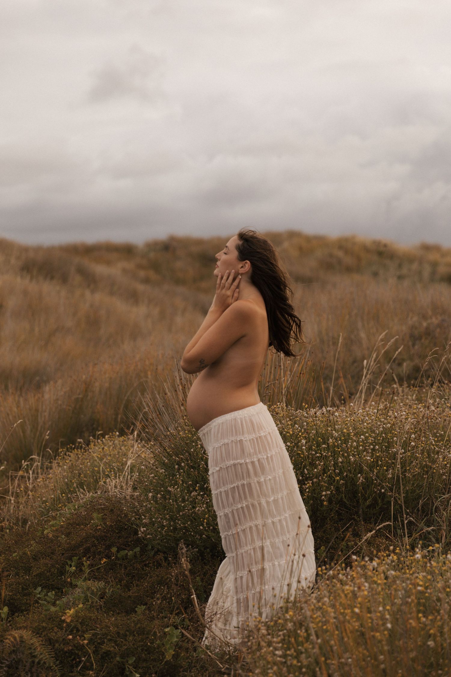 Artistic maternity photo shoot in a field of wildflowers at sunset with a white tiered dress.