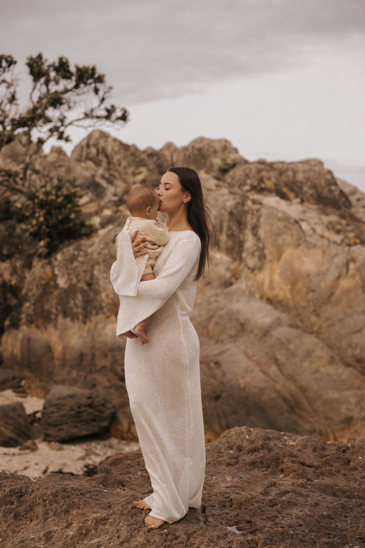 Artistic maternal photoshoot on rugged coastal terrain with muted earth tones.