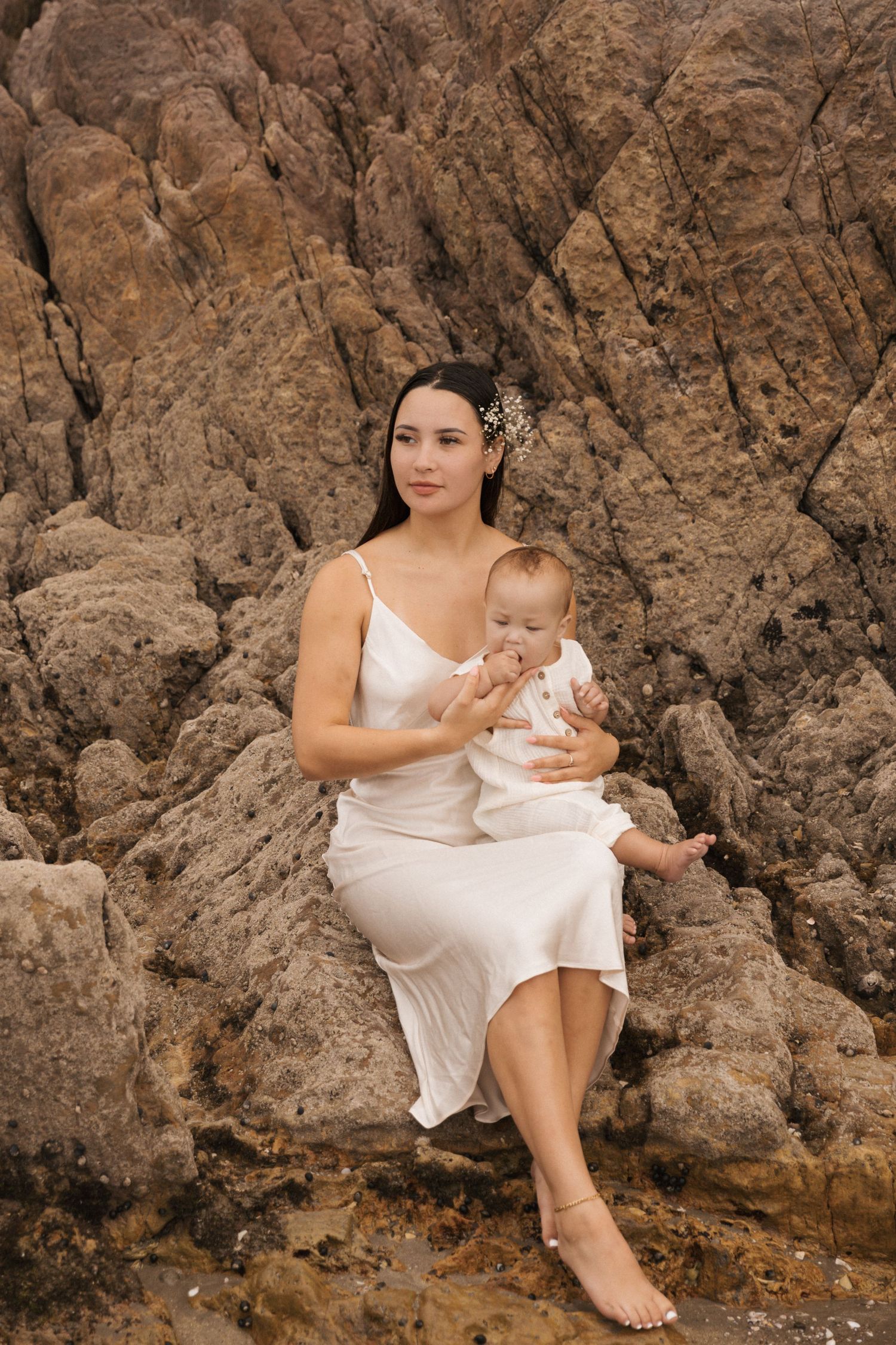 Mother and baby in matching white outfits posed against rocky coastal backdrop.